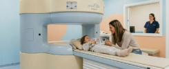 Fotografia de uma criança deitada confortavelmente na mesa de exame de uma máquina de ressonância magnética aberta Hitachi Airis Elite, segurando um brinquedo de pelúcia, enquanto sua mãe sorri para ela e segura sua mão. Uma técnica de radiologia observa da janela de controle. O ambiente é acolhedor, com paredes coloridas em azul e pêssego.